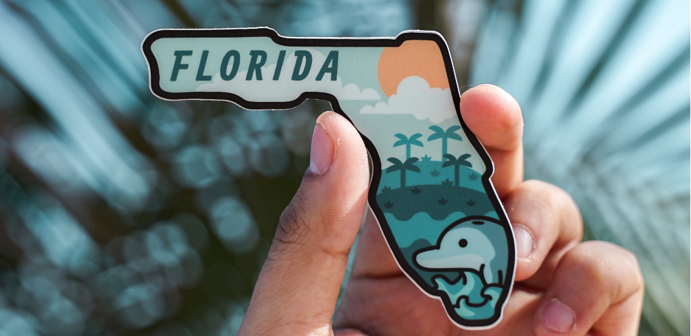 A magnet in the shape of the State of Florida with a cartoon dolphin, palm trees, clouds, sun and the word FLORIDA printed on the front is held up in the air by one hand with short nails on a blurred background.